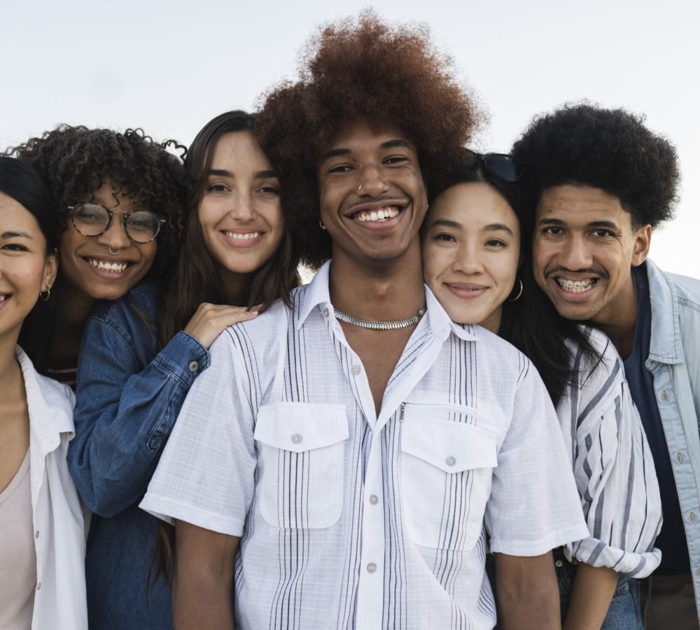 A close-up of a diverse group of young adults smiling and embracing each other outdoors. They appear happy and connected.